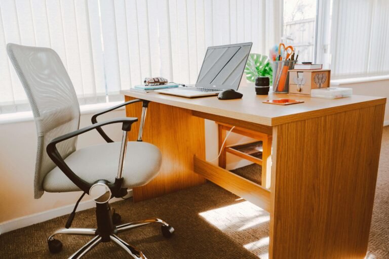 Cozy home office setup with a wooden desk, ergonomic chair, and a laptop, complemented by organized stationery and natural light filtering through vertical blinds.