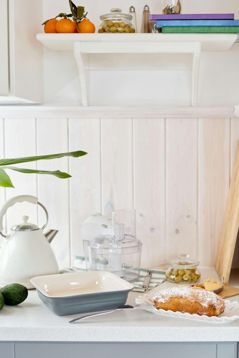 A bright kitchen counter with a white kettle, a food processor, a plate of freshly baked cake dusted with powdered sugar, and a bowl of green olives. A shelf above holds oranges and kitchen essentials.