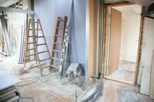 Interior of a home under renovation, showing drywall installation in progress. Two wooden ladders are leaning against a partially finished wall, with various construction materials and tools scattered around the dusty floor. A doorway to another room is visible on the right.