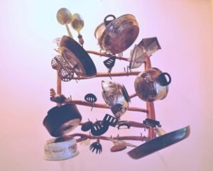 Vintage kitchen utensils and cookware arranged in a sculptural formation, including pots, pans, colanders, and various cooking tools, against a pink gradient background