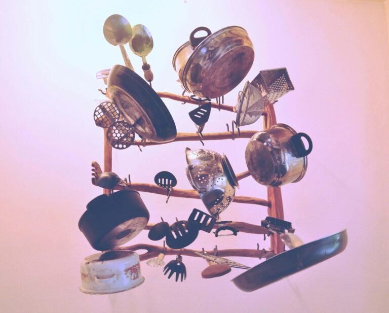 Vintage kitchen utensils and cookware arranged in a sculptural formation, including pots, pans, colanders, and various cooking tools, against a pink gradient background