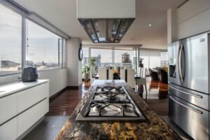Modern luxury open-plan kitchen with a large granite island featuring a gas cooktop, stainless steel range hood, and panoramic city views through floor-to-ceiling windows. The space includes white cabinetry, hardwood floors, and flows into a dining area with contemporary furniture.