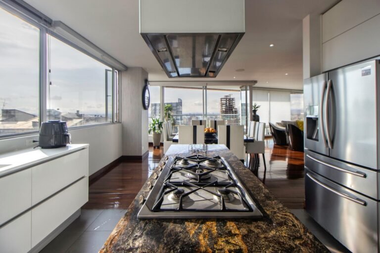 Modern luxury open-plan kitchen with a large granite island featuring a gas cooktop, stainless steel range hood, and panoramic city views through floor-to-ceiling windows. The space includes white cabinetry, hardwood floors, and flows into a dining area with contemporary furniture.