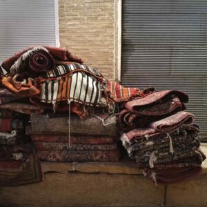 Stacked vintage rugs and carpets in rich reds and earth tones, piled against a brick wall with metal shutters. The textiles show traditional patterns and some wear, suggesting authenticity and age