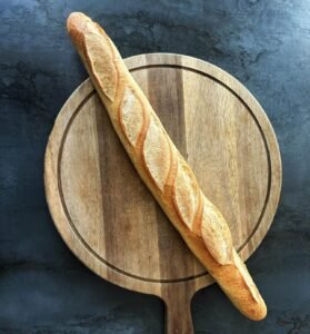 Freshly baked golden French baguette placed diagonally across a round wooden cutting board on a dark textured surface