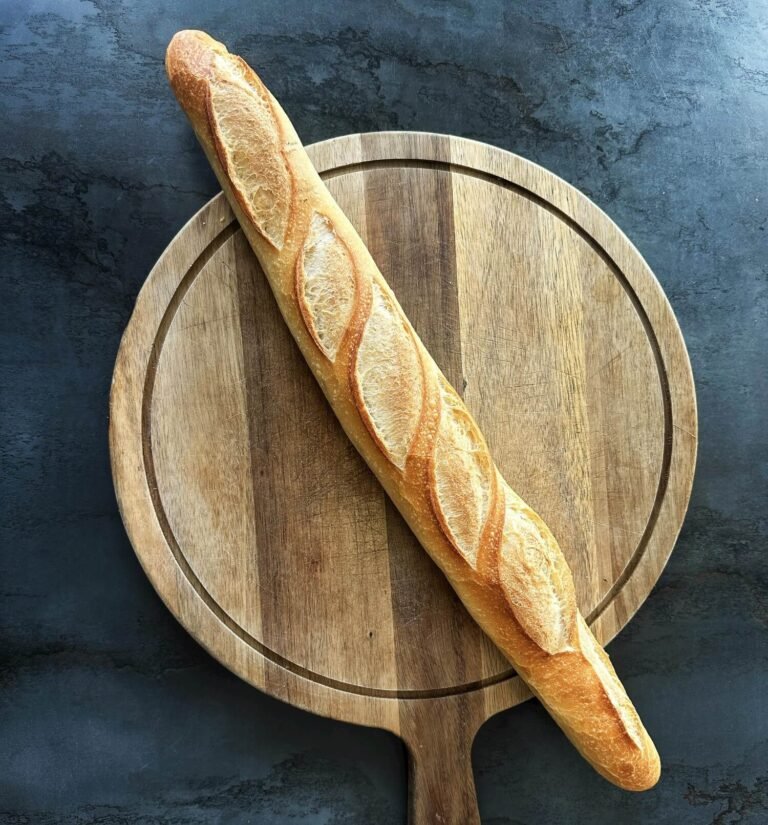 Freshly baked golden French baguette placed diagonally across a round wooden cutting board on a dark textured surface