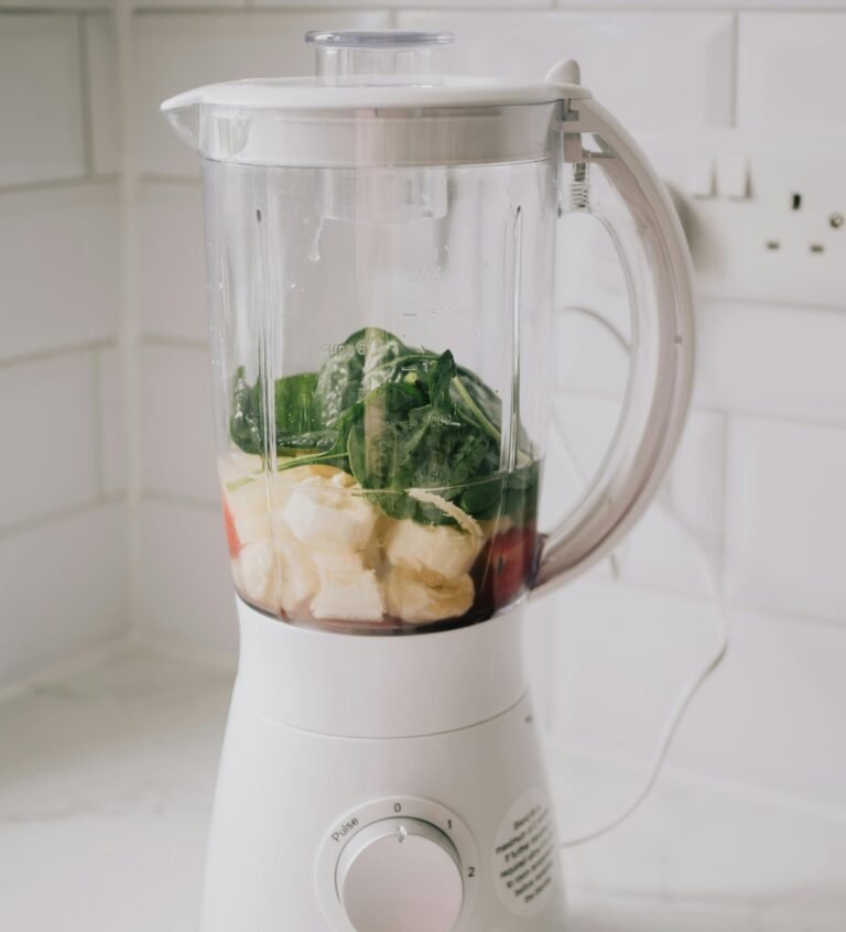 White blender filled with fresh ingredients for a smoothie, including bananas, spinach, and red fruit, sitting on a kitchen counter