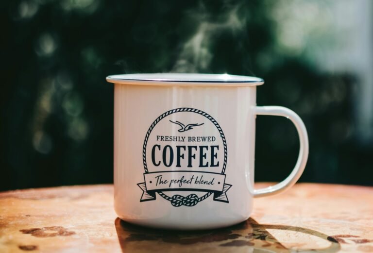 A white enamel mug with "Freshly Brewed Coffee: The perfect blend" logo sits on a wooden surface. The rustic mug design evokes a cozy, outdoorsy feeling against a blurred green background.