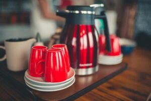 Coffee service featuring a red and black thermal carafe with matching bright red mugs on saucers, arranged on a wooden surface in a kitchen setting