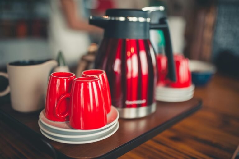 Coffee service featuring a red and black thermal carafe with matching bright red mugs on saucers, arranged on a wooden surface in a kitchen setting