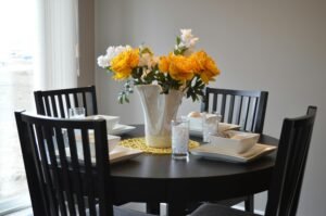 An elegantly set black dining table with four chairs, featuring a white vase filled with vibrant yellow roses and white flowers as a centerpiece. The table is set with white plates, glasses, and a yellow doily, creating a sophisticated yet cozy dining atmosphere near a window.