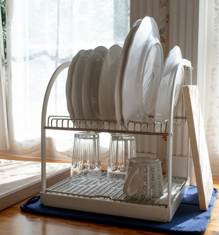 A white metal dish drying rack near a sunny window, filled with clean white plates, upside-down glass tumblers, and a decorative mug. The rack sits on a blue mat on a wooden surface, with curtains visible in the background.