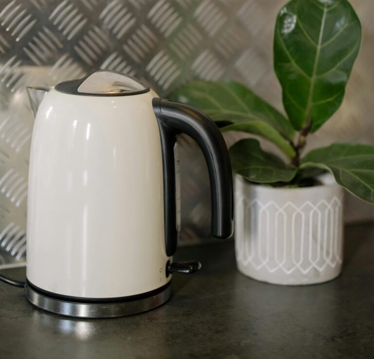 A sleek white electric kettle with black accents sits on a dark countertop, next to a white geometric-patterned plant pot containing a fiddle leaf fig plant. The background features a metallic patterned surface, creating a modern and stylish kitchen aesthetic.