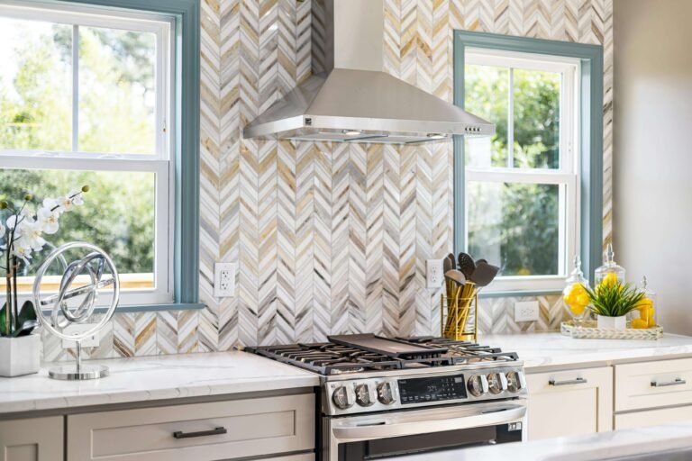 Modern kitchen featuring a striking chevron-patterned tile backsplash in white and gold tones, stainless steel gas range and hood, white countertops, and teal-framed windows offering natural light.