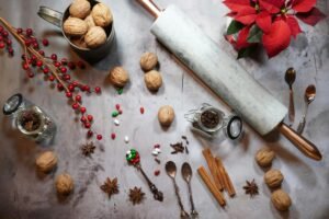Festive baking flatlay featuring a marble rolling pin, walnuts, cinnamon sticks, star anise, red berries, poinsettia, spices, and decorative candies arranged on a light surface