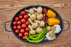An assortment of fresh vegetables including cherry tomatoes, mushrooms, bell peppers, green chilies, garlic, and dried red chilies arranged in a black cast iron pan on a wooden surface.
