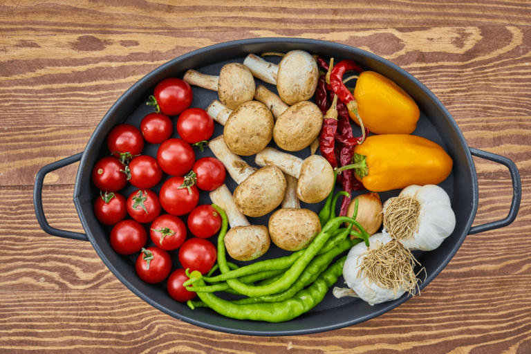 An assortment of fresh vegetables including cherry tomatoes, mushrooms, bell peppers, green chilies, garlic, and dried red chilies arranged in a black cast iron pan on a wooden surface.