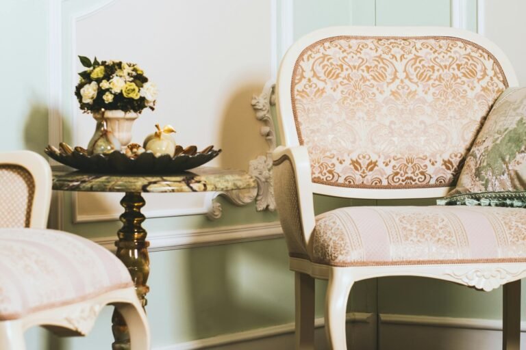 Ornate vintage living room featuring a cream-colored armchair with intricate floral upholstery, an antique side table with a decorative vase of yellow roses, and classic wall moldings