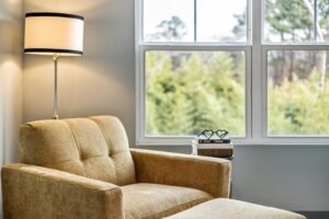 Comfortable beige armchair with ottoman in a bright corner, next to a window overlooking green foliage. A table lamp and stacked books with a heart-shaped decoration complete the cozy reading nook.