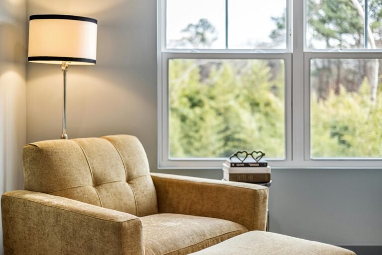 Comfortable beige armchair with ottoman in a bright corner, next to a window overlooking green foliage. A table lamp and stacked books with a heart-shaped decoration complete the cozy reading nook.
