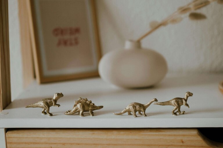 Four small gold dinosaur figurines arranged on a white shelf, with a white vase and framed artwork in the background, creating a stylish and whimsical home decor display.