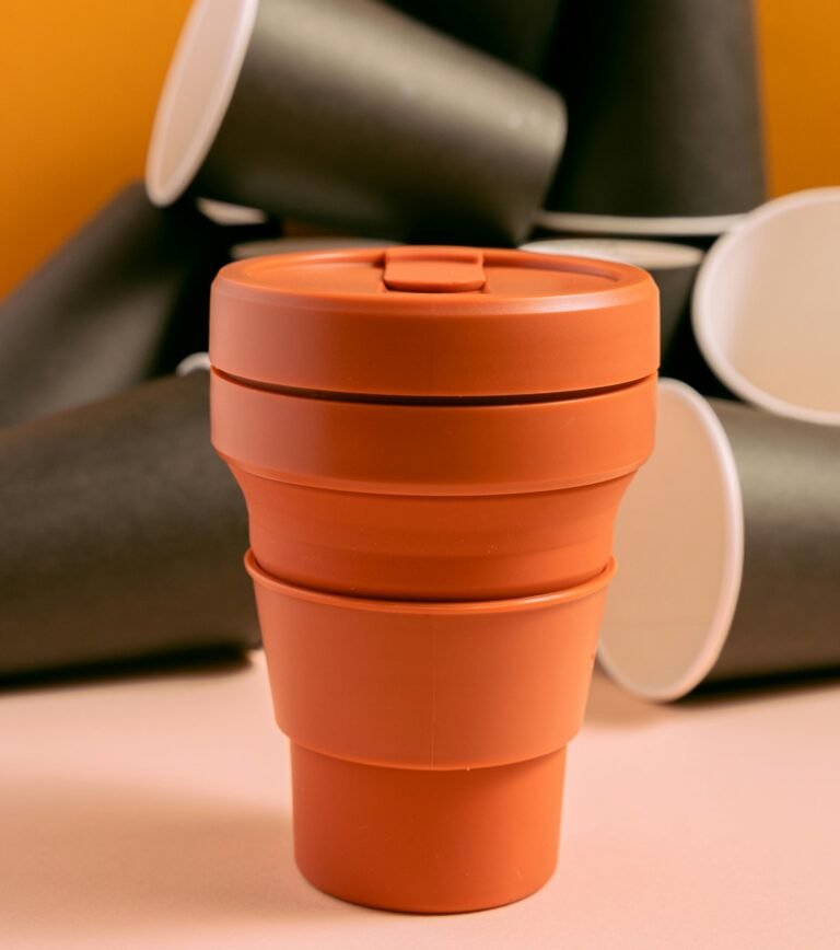 Orange collapsible silicone coffee cup with lid, shown expanded on a table with other cups visible in the background