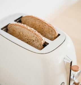 Two slices of whole grain bread toasting in a sleek white toaster. The bread has a golden crust with visible grains on top, sitting in a modern appliance with minimalist design and rose gold accents.