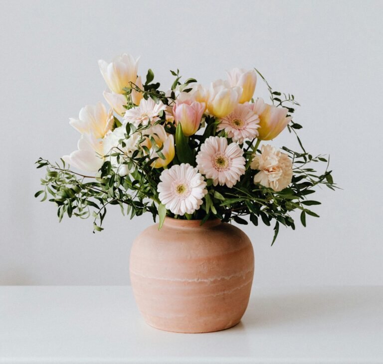 Elegant spring floral arrangement featuring pale pink gerberas, yellow tulips, and lush greenery in a rustic terracotta vase against a light background.