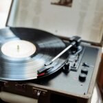 Close-up of a vintage record player with a vinyl record spinning. The tonearm and stylus are visible on the black disc. The turntable has a textured case and control knobs, capturing retro audio nostalgia.