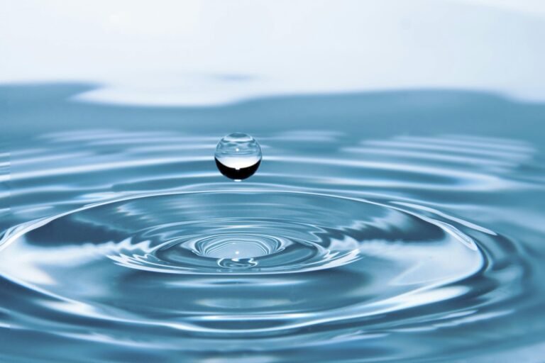 Close-up macro photograph of a pristine water droplet hovering above a calm water surface. The image captures concentric circular ripples and a perfectly spherical water drop, showcasing the clarity and purity of water while demonstrating surface tension and fluid dynamics.