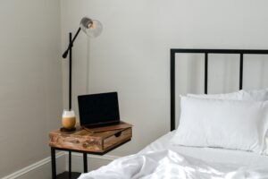 Minimalist bedroom with black metal bed, wooden bedside table, open laptop, and latte under a wall lamp.